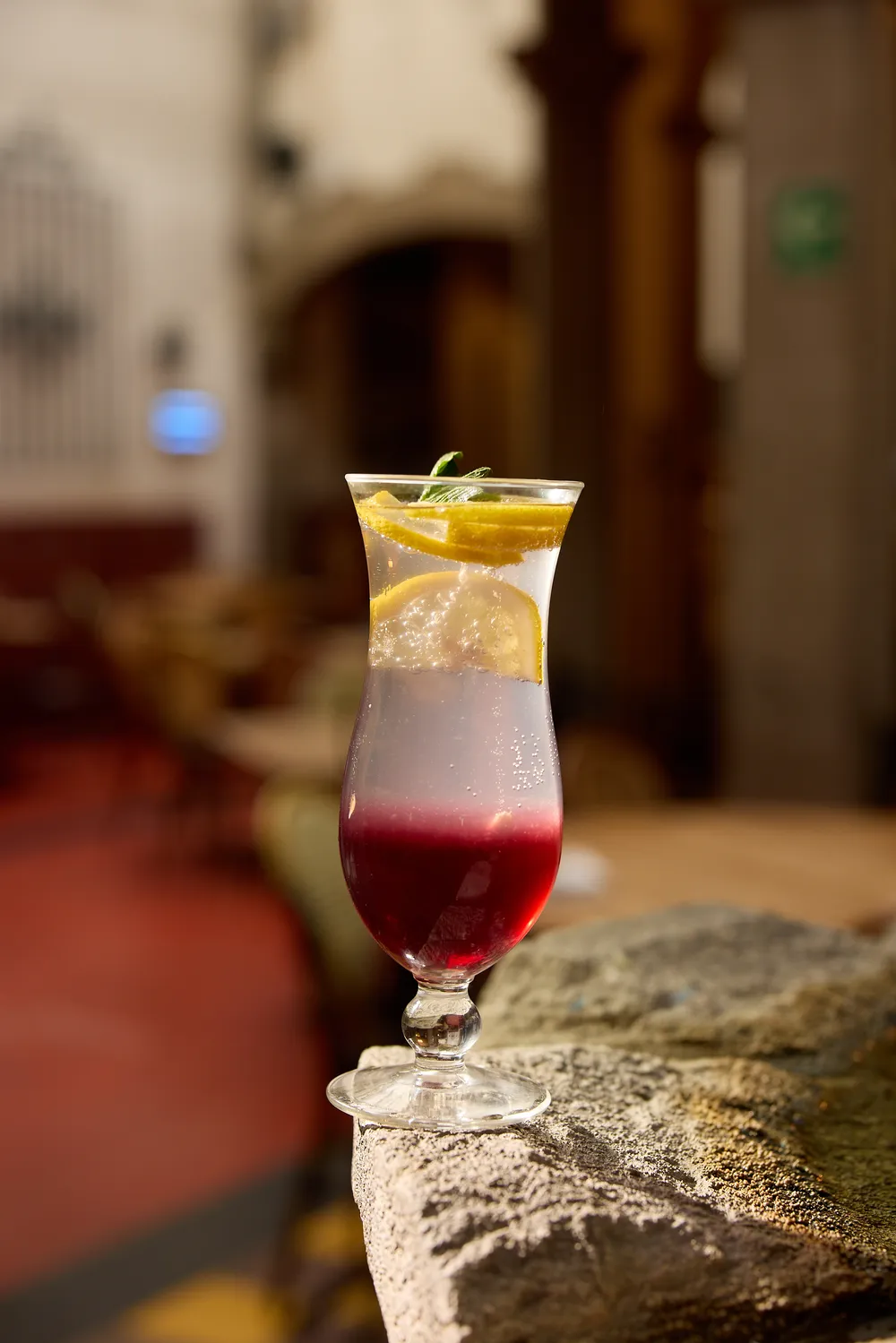 Coctel de capas rojas y amarillas con hielo y romero sobre una barda de piedra, con el interior del restaurante desenfocado al fondo