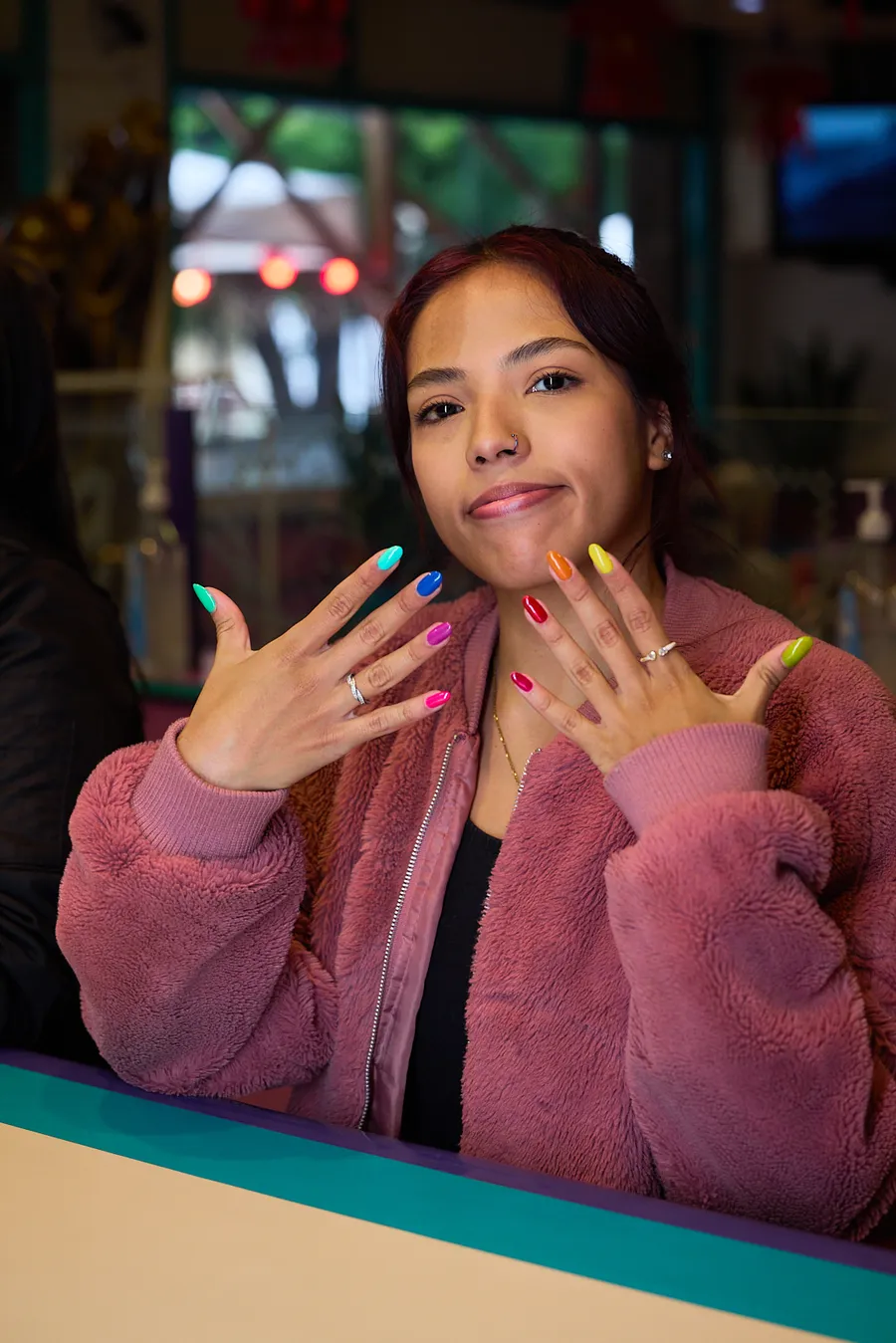 Retrato en locación: joven con abrigo rosa mostrando a cámara las uñas pintadas de colores
