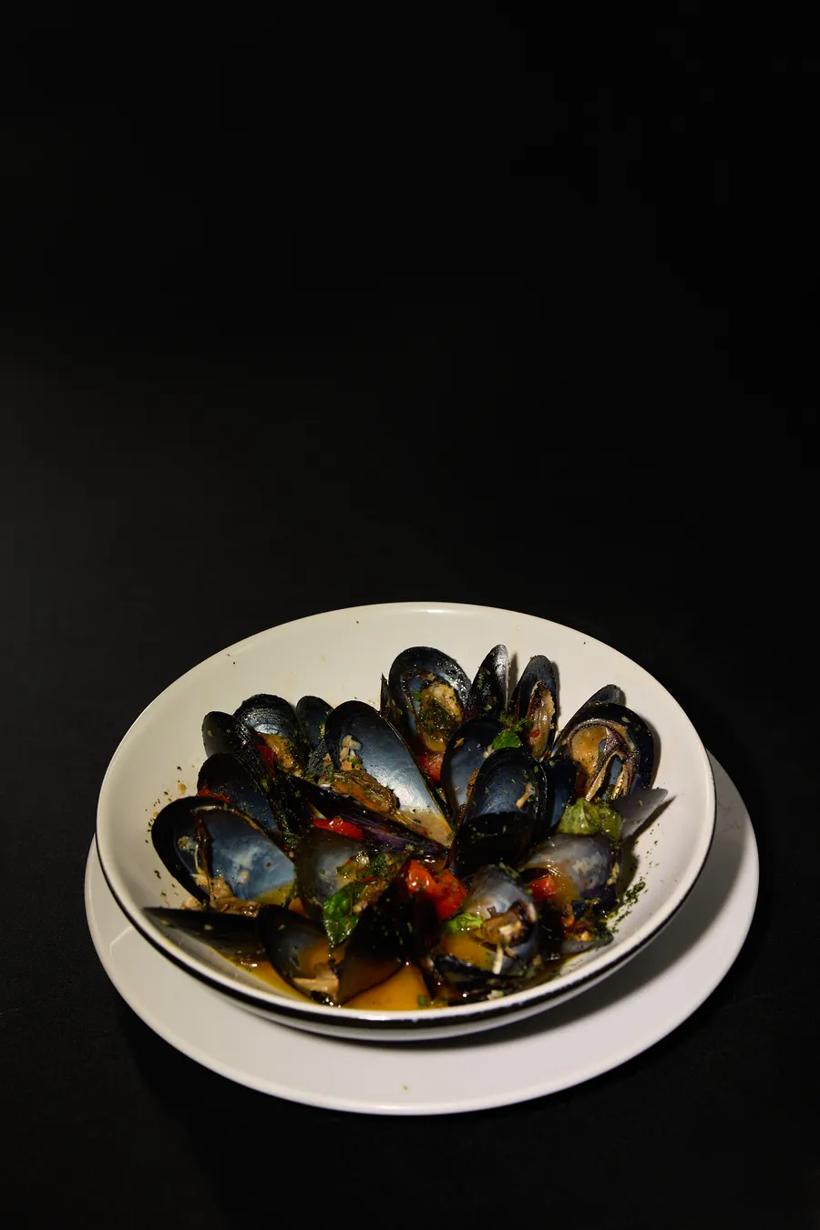 Plato de mejillones al vapor con jitomate y perejil, sobre vajilla blanca y fondo negro absoluto