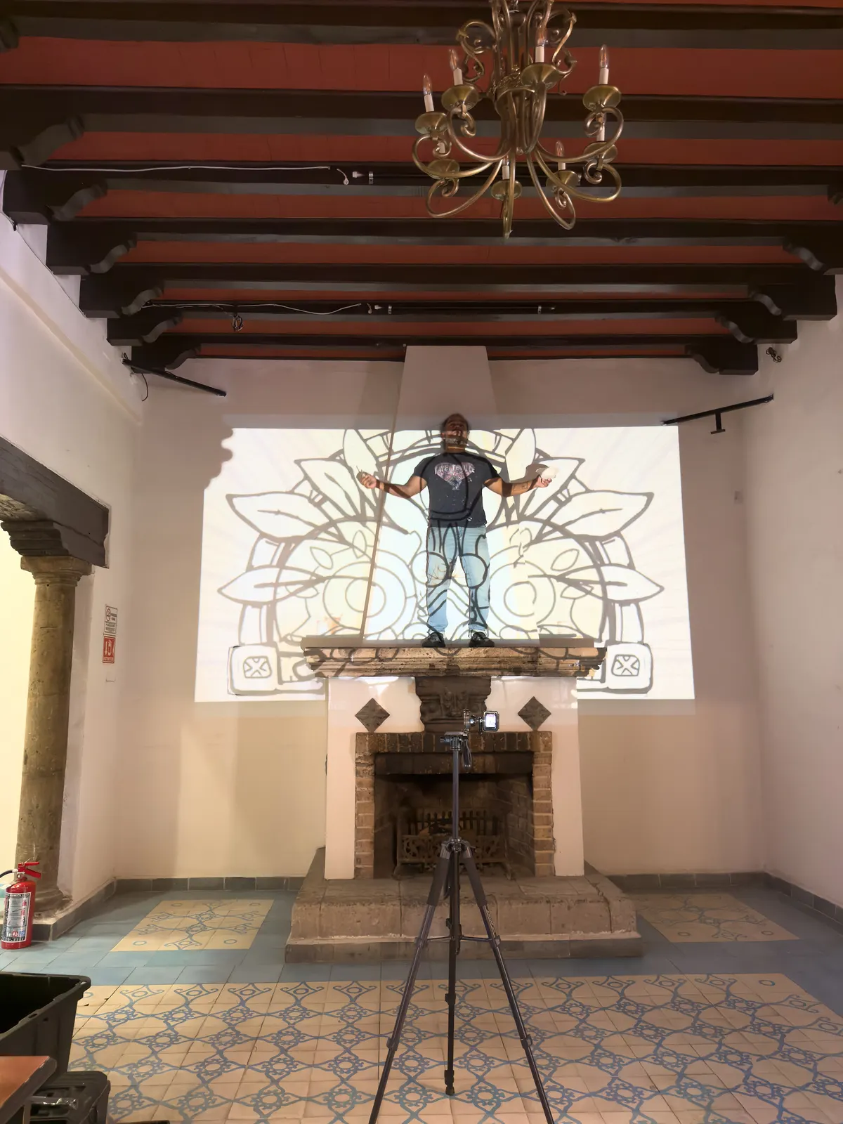Pintor de pie sobre la chimenea de un restaurante trazando un mural con el diseño proyectado en la pared, mientras una cámara en tripié graba el proceso