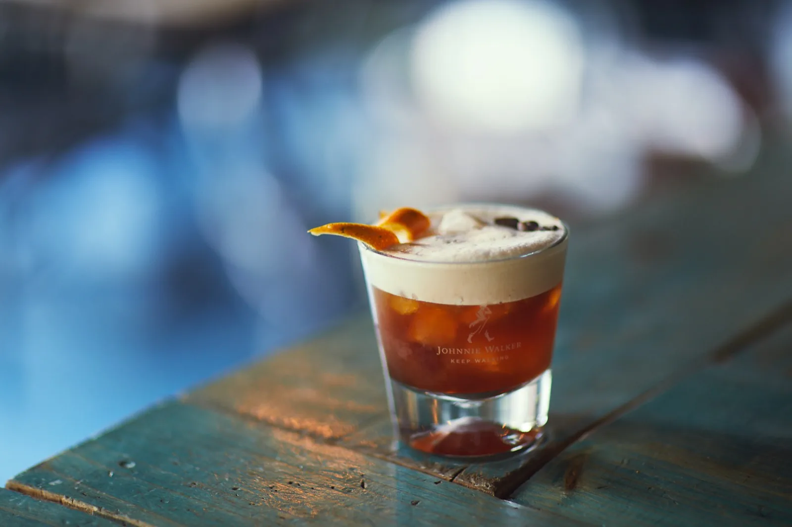 Coctel servido en Cabo Coyote: vaso corto con espuma y cáscara de naranja tostada, sobre una mesa de madera turquesa